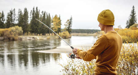 Young man fisherman in a yellow sweater and hat is fishing on the river bank in the autumn forest with a spinning rod in his hands. Concept of relaxing outdoor recreation, lifestyle, hobbyの写真素材