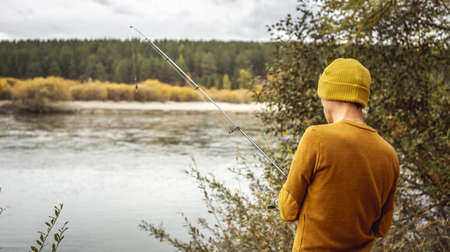 Young man fisherman in a yellow sweater and hat is fishing on the river bank in the autumn forest with a spinning rod in his hands. Concept of relaxing outdoor recreation, lifestyle, hobbyの写真素材