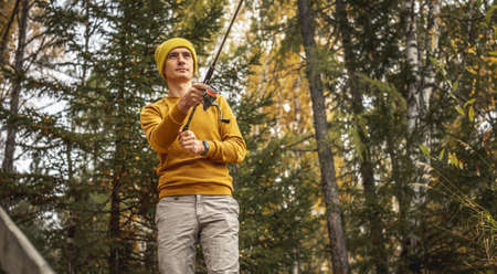 Young man fisherman in a yellow sweater and hat is fishing on the river bank in the autumn forest with a spinning rod in his hands. Concept of relaxing outdoor recreation, lifestyle, hobbyの写真素材
