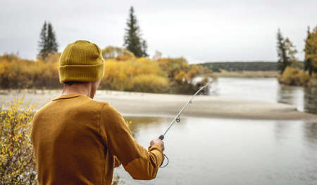 Young man fisherman in a yellow sweater and hat is fishing on the river bank in the autumn forest with a spinning rod in his hands. Concept of relaxing outdoor recreation, lifestyle, hobbyの写真素材