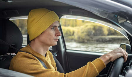 Man in yellow hat and sweater is driving a car along the river and the beautiful autumn forest. Concept of nature, travel and autumn moodの写真素材