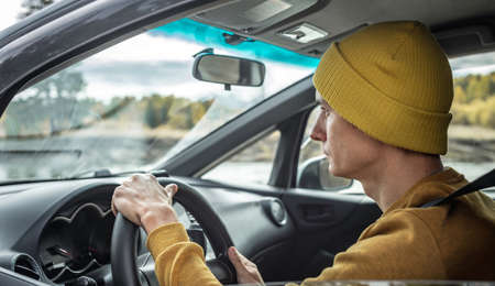 Man in yellow hat and sweater is driving a car along the river and the beautiful autumn forest. Concept of nature, travel and autumn moodの写真素材