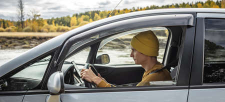 Man in yellow hat and sweater is driving a car along the river and the beautiful autumn forest. Concept of nature, travel and autumn moodの写真素材