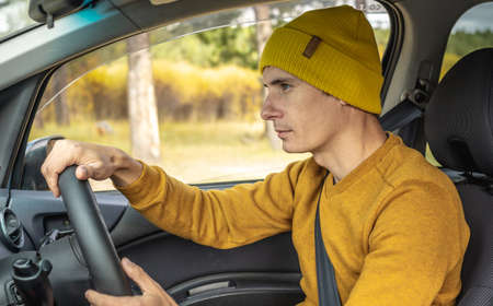 Man in yellow hat and sweater is driving a car along the autumn forest road. Concept of nature, travel and autumn mood.の写真素材