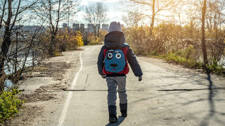 A child boy hiking with a small backpack is walking along the road outside the city on the background of the autumn landscapeの写真素材