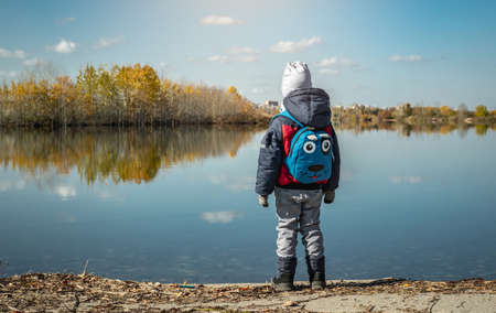 A child boy hiking with a backpack stands on the shore of the lake and looks at the reflection of the blue skyの写真素材