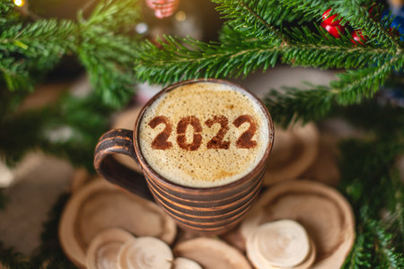 A mug of cappuccino coffee with numbers 2022 on a wooden background with green branches of a Christmas tree. Warm and cozy from a hot drink on Christmas morning.の写真素材