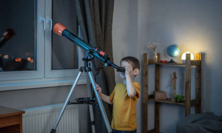 Cute boy is looking through a telescope in a room at the night starry sky. Children's scientific hobbies and space explorationの写真素材