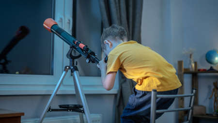 Cute boy is looking through a telescope in a room at the night starry sky. Children's scientific hobbies and space explorationの写真素材