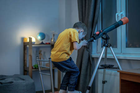 Cute boy is looking through a telescope in a room at the night starry sky. Children's scientific hobbies and space explorationの写真素材