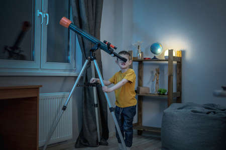 Cute boy is looking through a telescope in a room at the night starry sky. Children's scientific hobbies and space explorationの写真素材