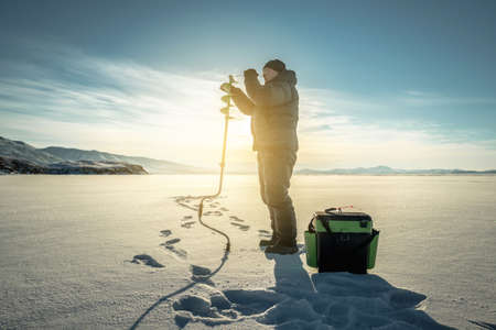 A fisherman drills a hole in the ice of a large frozen lake on a sunny day. The joy of winter fishingの写真素材
