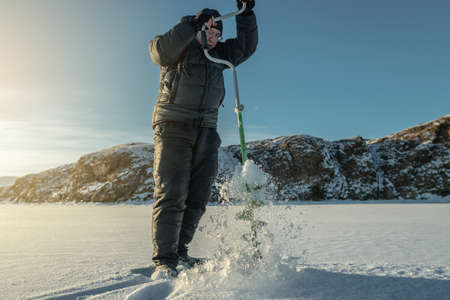 A fisherman drills a hole in the ice of a large frozen lake on a sunny day. The joy of winter fishingの写真素材
