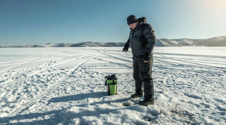 A fisherman is fishing in a hole on the ice of a large frozen lake on a sunny day. The joy of winter fishing on Lake Baikalの写真素材