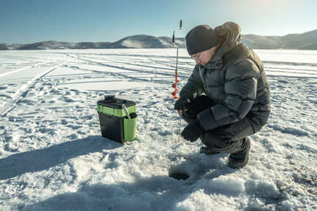 A fisherman is fishing in a hole on the ice of a large frozen lake on a sunny day. The joy of winter fishing on Lake Baikalの写真素材
