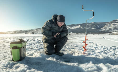 A fisherman is fishing in a hole on the ice of a large frozen lake on a sunny day. The joy of winter fishing on Lake Baikalの写真素材