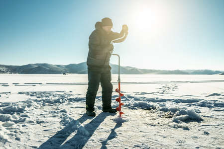 A fisherman drills a hole in the ice of a large frozen lake on a sunny day. The joy of winter fishingの写真素材