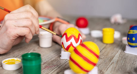 Woman's hand with a brush is drawing a colored pattern on an Easter egg. Preparation for the bright...
