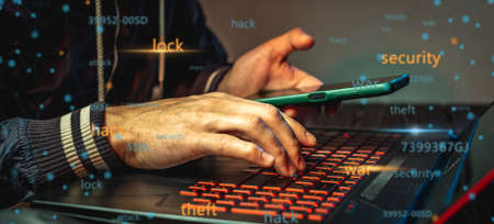 A hacker with a phone is typing with his hands on a laptop keyboard in a dark room. The concept of cyber warfare and Dos attacksの写真素材