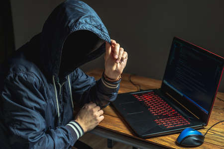 A hacker anonymous in a hood is typing on a laptop keyboard in a dark room. The concept of cybercrime fraud and identity theftの写真素材