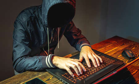 A hacker anonymous in a hood is typing on a laptop keyboard in a dark room. The concept of cyber warfare and Dos attacksの写真素材