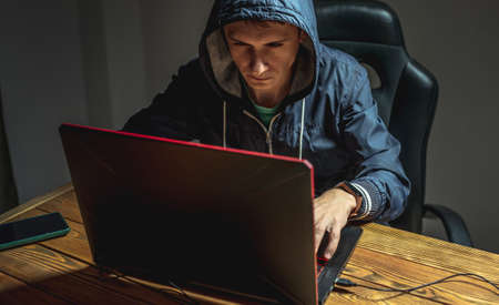 A hacker in a hood with a phone is typing on a laptop keyboard in a dark room. The concept of cybercrime fraud and identity theftの写真素材