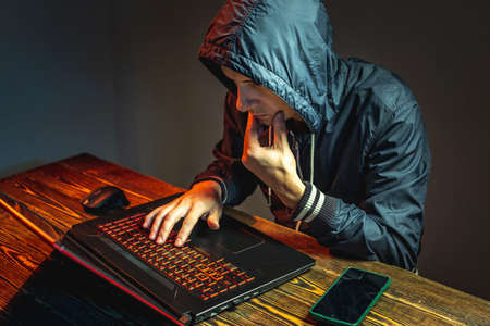 A hacker in a hood with a phone is typing on a laptop keyboard in a dark room. The concept of cyber warfare and Dos attacksの写真素材
