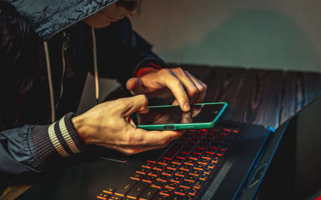 A hacker with a phone is typing with his hands on a laptop keyboard in a dark room. The concept of cyber warfare and Dos attacksの写真素材
