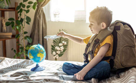A child boy with a large backpack is playing with a toy airplane and a globe of the earth. The concept of going on a journey towards adventure.の写真素材