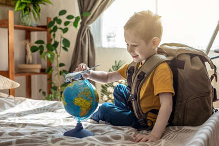A child boy with a large backpack is playing with a toy airplane and a globe of the earth. The concept of going on a journey towards adventure.の写真素材
