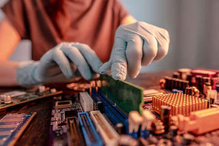 The engineer's gloved hand holds the computer's RAM chip against the background of the motherboard. The concept of high-tech hardware microelectronicsの写真素材