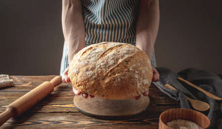 Female hands holding homemade natural fresh bread with a Golden crust on a napkin on an old wooden background . The concept of baking bakery products. rustic style.の写真素材