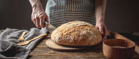 Women's hands cut with a knife homemade natural fresh bread with a Golden crust on a napkin on an old wooden background. The concept of baking bakery productsの写真素材