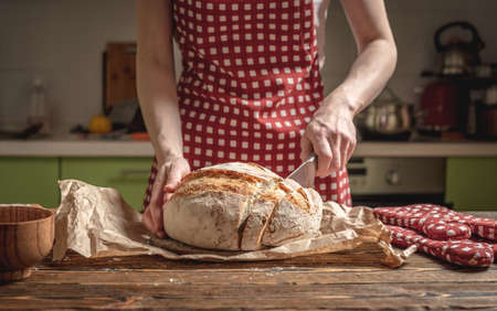 Women's hands cut with a knife homemade natural fresh bread with a Golden crust on a napkin on an old wooden background. The concept of baking bakery productsの写真素材
