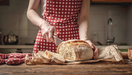 Women's hands cut with a knife homemade natural fresh bread with a Golden crust on a napkin on an old wooden background. The concept of baking bakery productsの写真素材