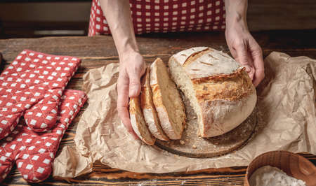 Women's hands cut with a knife homemade natural fresh bread with a Golden crust on a napkin on an old wooden background. The concept of baking bakery productsの写真素材