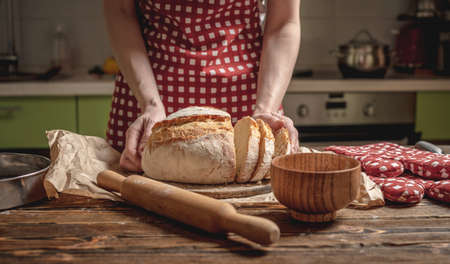 Women's hands cut with a knife homemade natural fresh bread with a Golden crust on a napkin on an old wooden background. The concept of baking bakery productsの写真素材