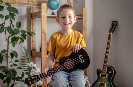 Cute funny boy is playing ukulele guitar in the music room. Joyful learning to play musical instruments.の写真素材