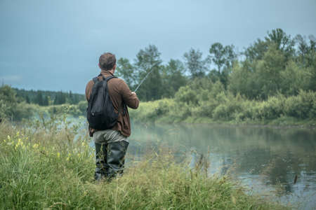 Male fisherman is standing on the riverbank with a fishing rod in his hands and catching fish. Concept of Summer calm outdoor recreation, pastime, hobbyの写真素材