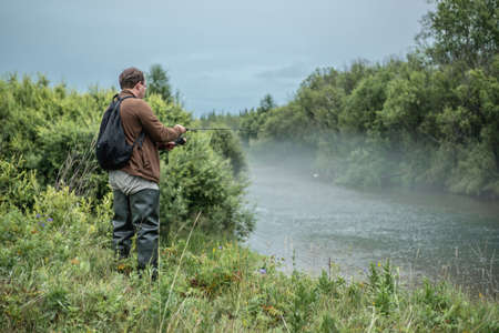 Male fisherman is standing on the riverbank with a fishing rod in his hands and catching fish. Concept of Summer calm outdoor recreation, pastime, hobbyの写真素材