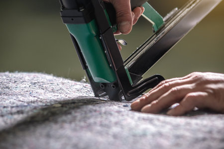 An employee holds in his hands an industrial air stapler for sheathe soft fabric upholstery. Close-up. Manufacture of upholstered furnitureの写真素材