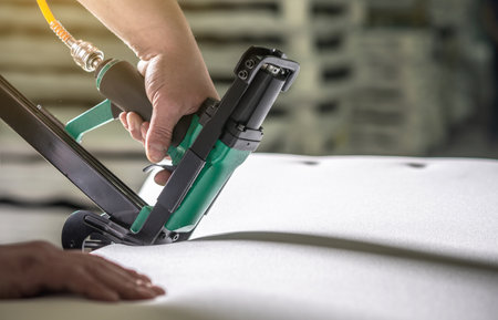 An employee holds in his hands an industrial air stapler for sheathe soft fabric upholstery. Close-up. Manufacture of upholstered furnitureの写真素材