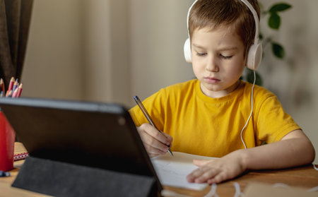 The child is a boy in a yellow T-shirt with headphones doing homework on a tablet. The concept of remote school education at home.の写真素材