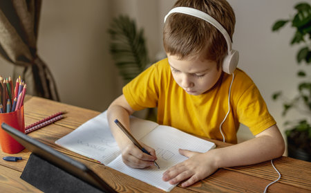 The child is a boy in a yellow T-shirt with headphones doing homework on a tablet. The concept of remote school education at home.の写真素材