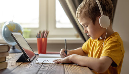 The child is a boy in a yellow T-shirt with headphones doing homework on a tablet. The concept of remote school education at home.の写真素材