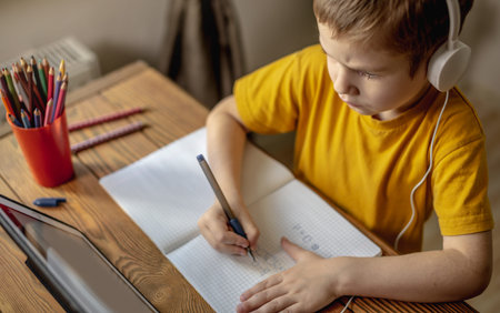 The child is a boy in a yellow T-shirt with headphones doing homework on a tablet. The concept of...