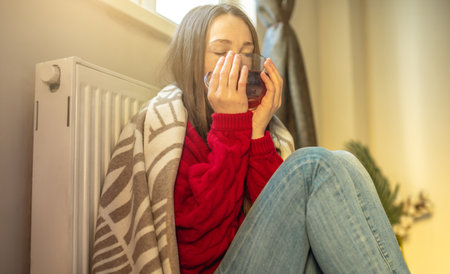 A woman wrapped in a plaid is sitting on the floor with hot tea and warming herself near a warm radiator. The concept of economy and the crisis of energy resources.の写真素材