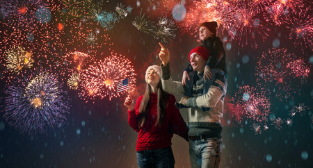 Mom and dad with their son on their shoulders in warm clothes and a USA flag in their hand. The family celebrates the New Year by looking at the fireworks outside. Happy Christmas weekend.の写真素材