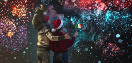 Mom and dad with their son on their shoulders in warm clothes and a italy flag in their hand. The family celebrates the New Year by looking at the fireworks outside. Happy Christmas weekend.の写真素材