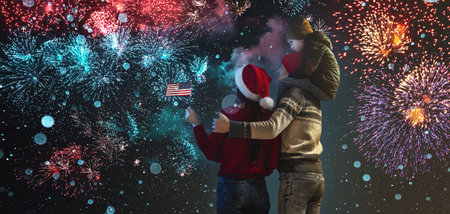 Mom and dad with their son on their shoulders in warm clothes and a USA flag in their hand. The family celebrates the New Year by looking at the fireworks outside. Happy Christmas weekend.の写真素材